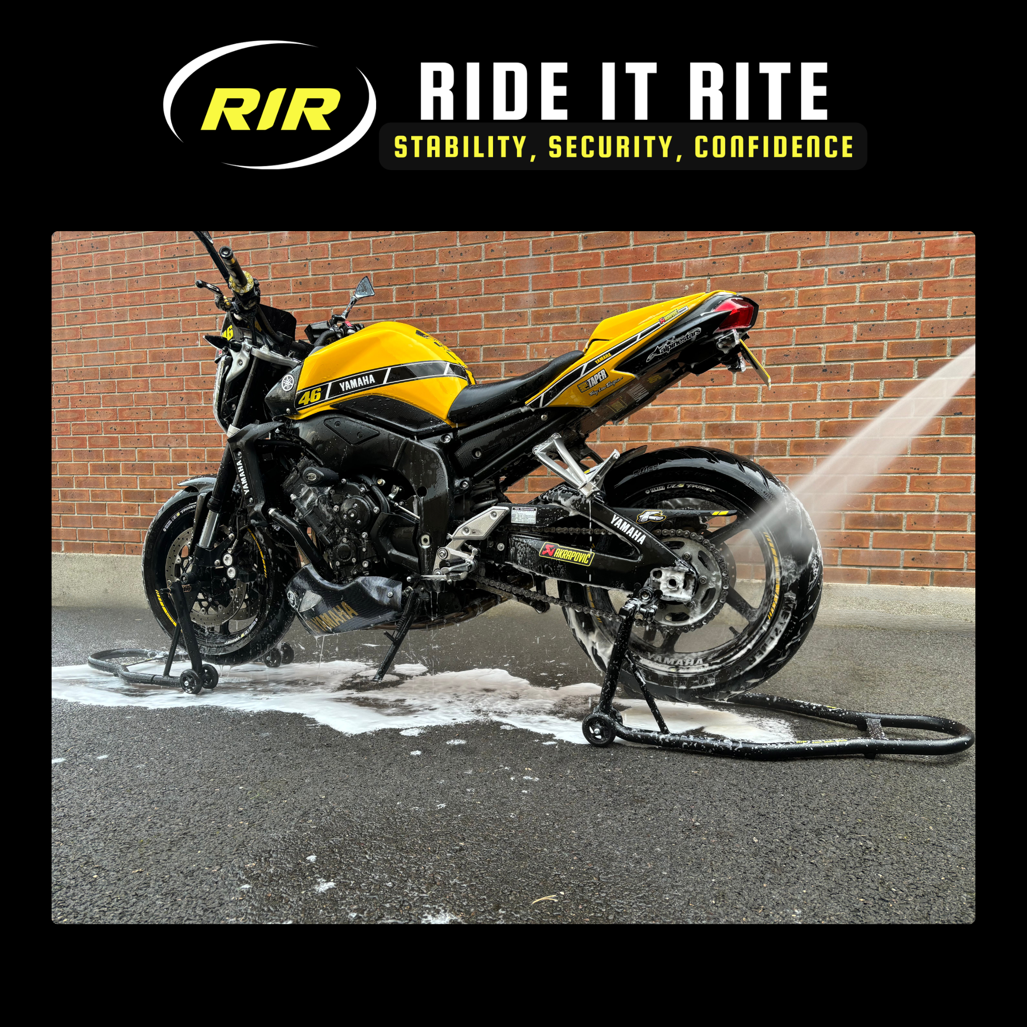 Yellow and black motorcycle being washed with a hose, sitting on 'Ride It Rite' black paddock stands with a brick wall background.