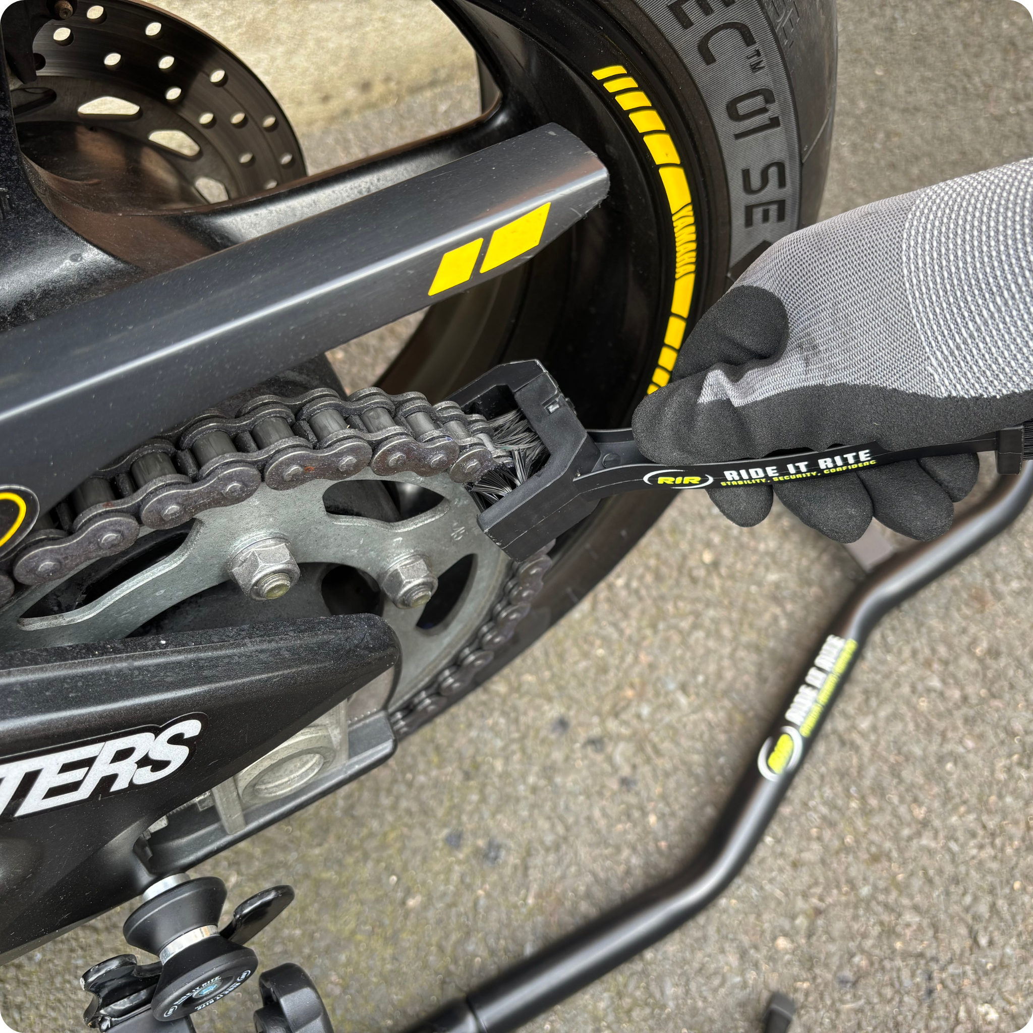 Close-up of a gloved hand cleaning a motorcycle chain with a black Ride It Rite chain cleaning brush.