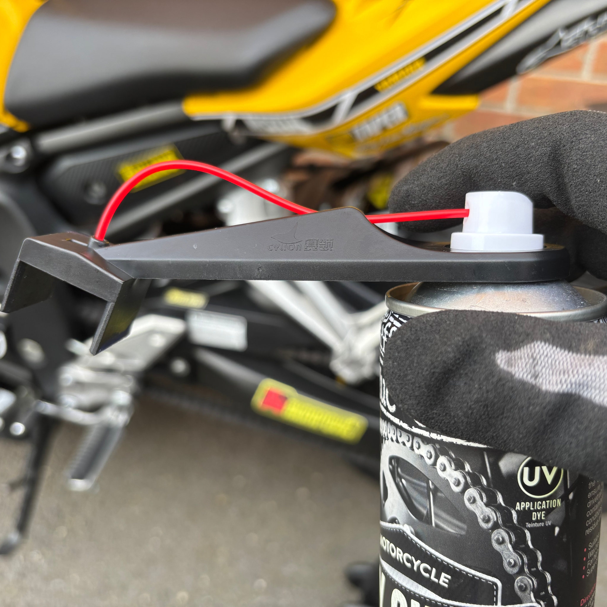 Motorcycle chain spray can with a Ride It Rite oil sprayer on it in a gloved hand holding it, with a motorcycle in the background.