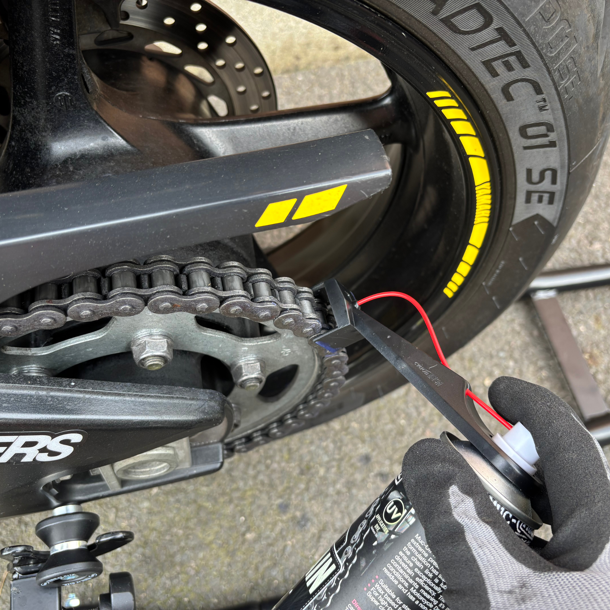 Close-up of a motorcycle chain with a gloved hand applying lubricant using a Ride It Rite oil sprayer, on a concrete surface.
