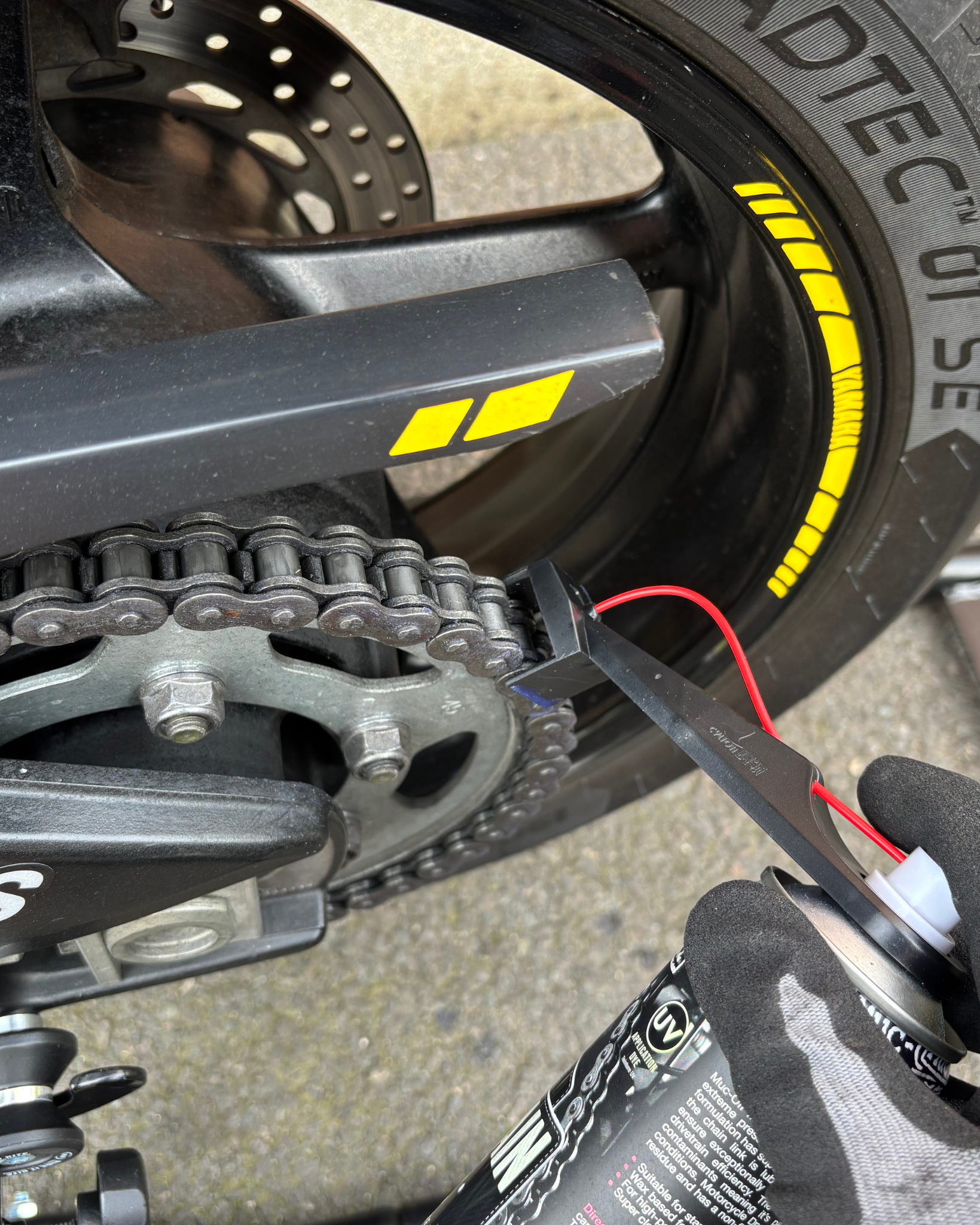 Close-up of a motorcycle chain with a gloved hand applying lubricant using a Ride It Rite oil sprayer, on a concrete surface.