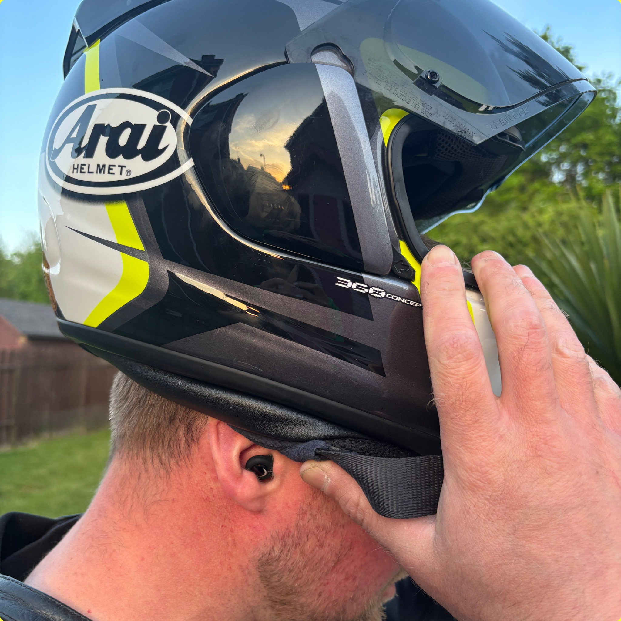 Person putting on a black and yellow Arai motorcycle helmet outdoors with Ride It Rite black ear plugs in ear.