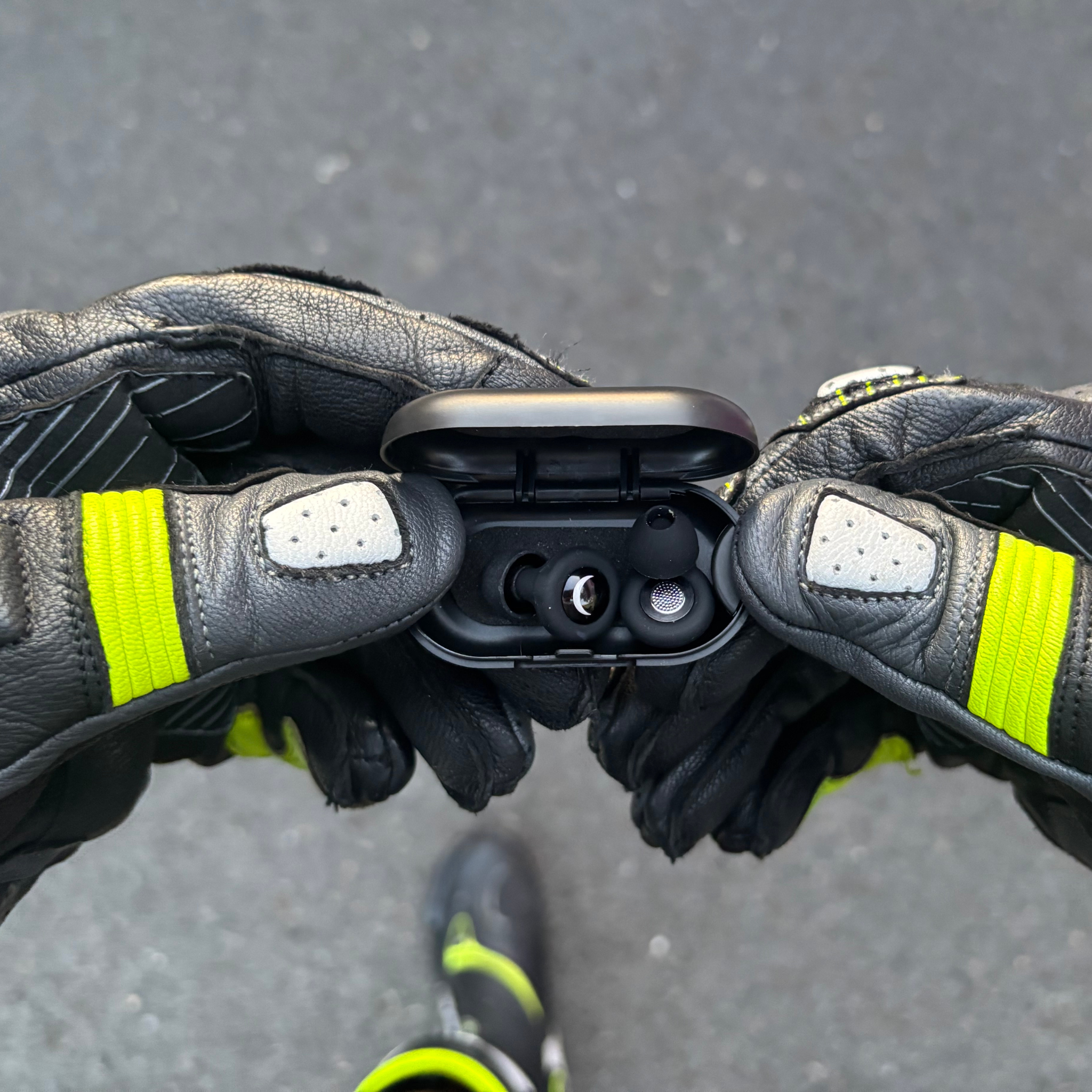 Close-up of hands wearing black gloves with yellow accents holding Ride It Rite black ear plugs in their case on a grey pavement background.