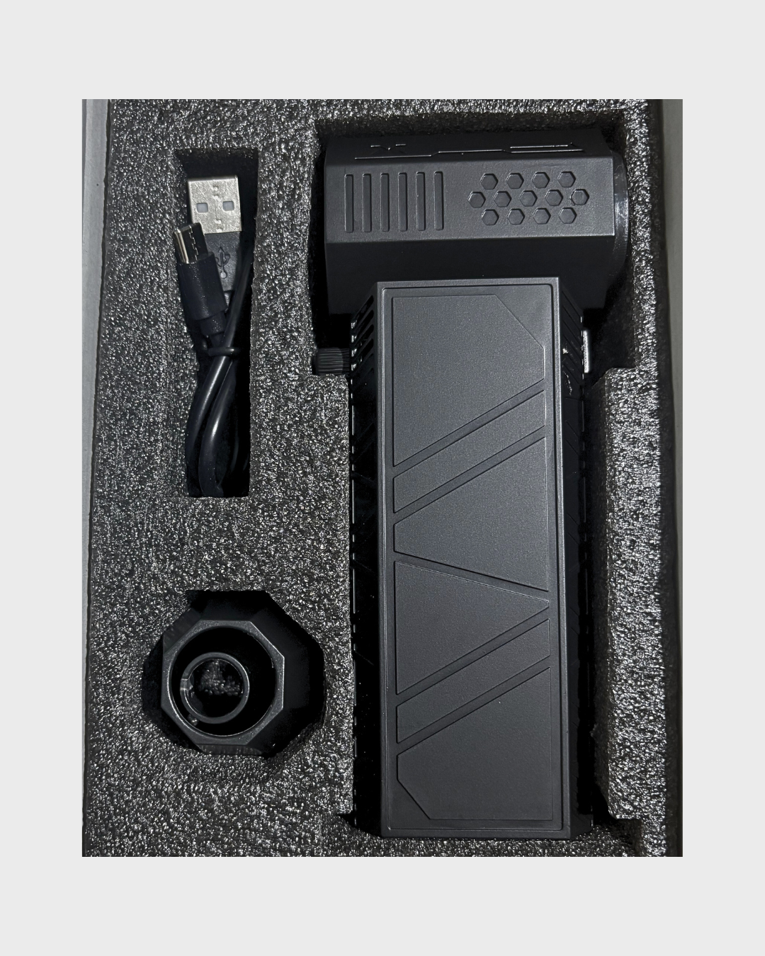 A black  handheld motorcycle air blower with a long rectangular body and angled panel details, shown inside a foam-lined presentation box alongside a USB cable and a round black accessory.