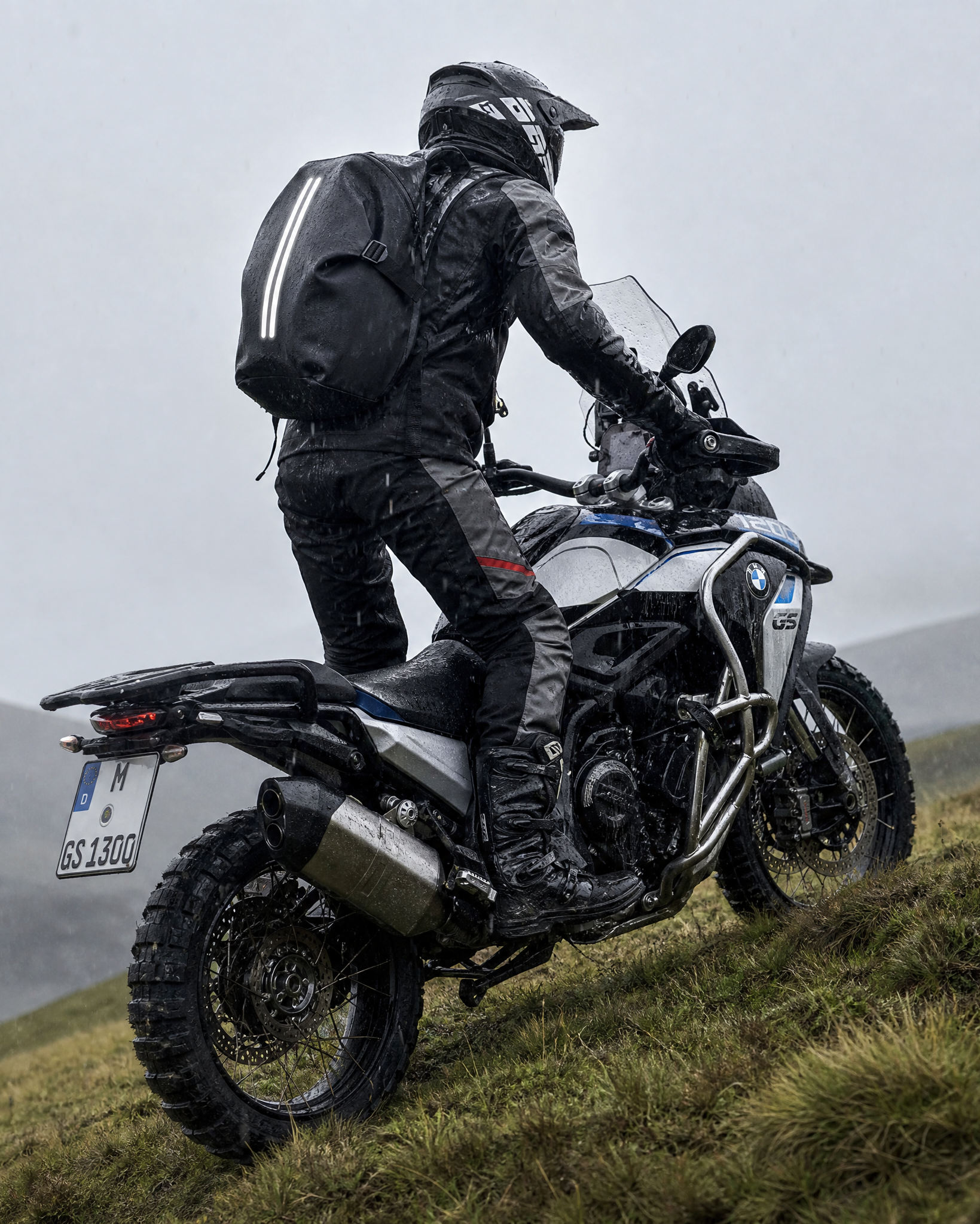 A rider in full black motorcycle gear wearing a waterproof motorcycle backpack and helmet sits on a white and blue BMW adventure bike, photographed from behind as they ride up a grassy hill under an overcast sky.