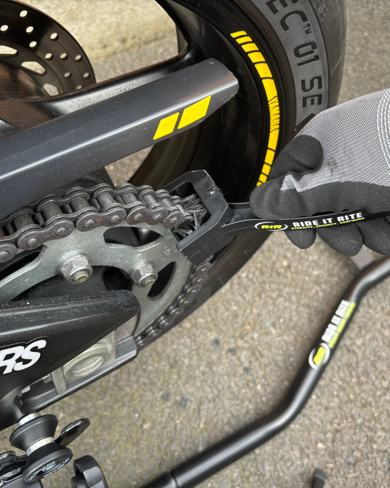 Close-up of a gloved hand cleaning  a motorcycle chain with a black Ride It Rite  chain cleaning brush.